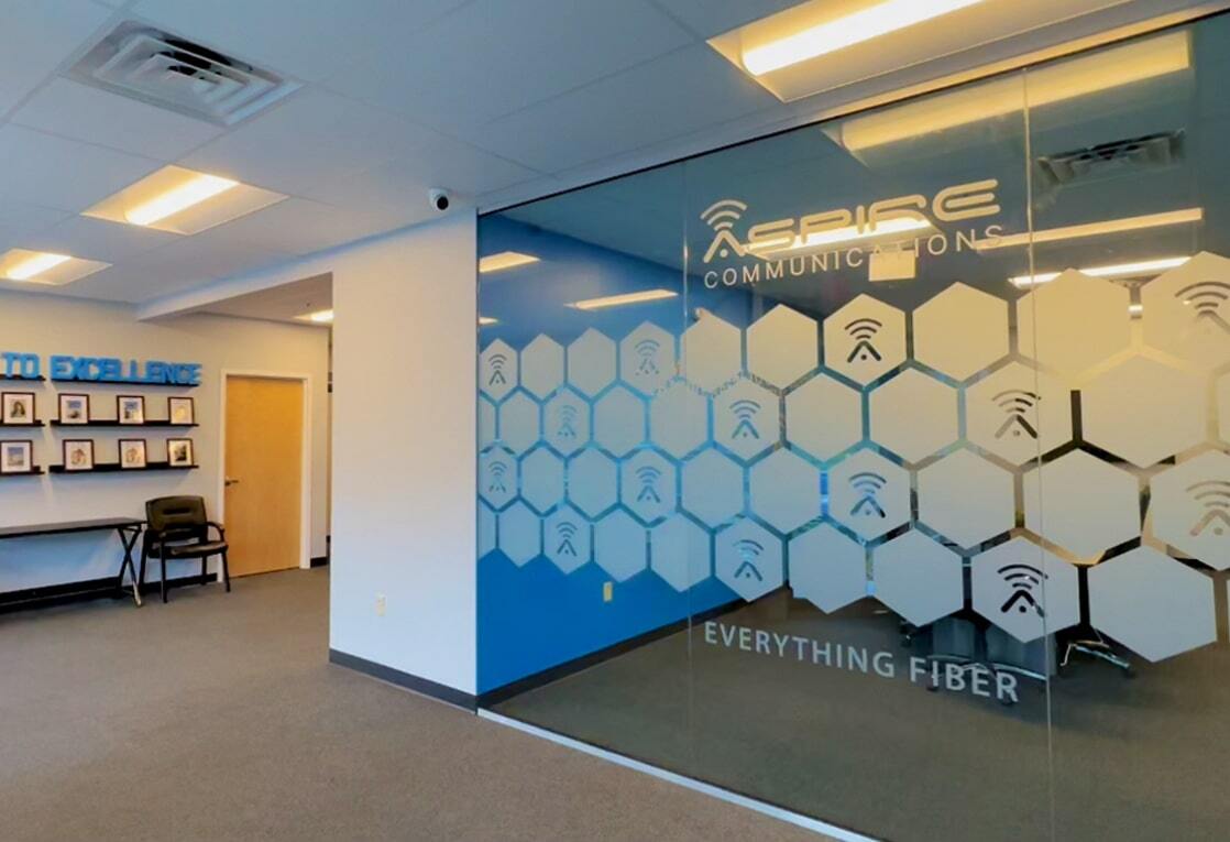 This is a photo of the Aspire Communications office. The office space has a glass wall and door. The glass wall has a blue and white hexagonal pattern on it. The door has a blue and white sign that reads “Aspire Communications”. The wall behind the door has a sign that reads “Aspire to Excel”. The wall to the right of the door has a sign that reads “Everything Fiber”. The office space has a desk and chairs, and a window with a view of trees.