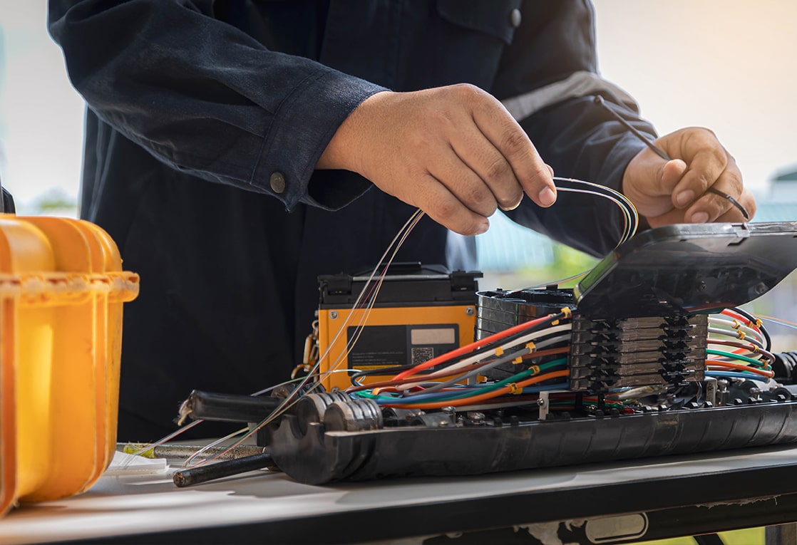 A person’s hands working on an electrical panel. The person is wearing a black jacket and is holding a wire in their right hand. The electrical panel is black and has multiple wires of different colors connected to it.